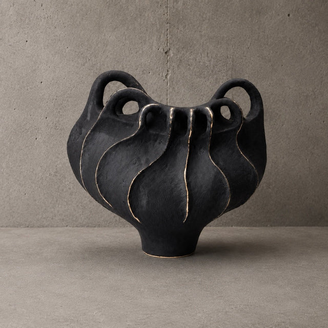 A black ceramic sculptural vase with three loop handles and a ribbed, wavy surface, displayed on a concrete surface against a textured concrete wall.