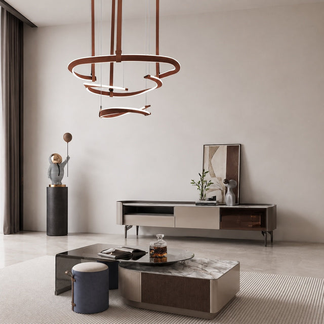 A modern living room interior featuring a sculptural multi-ring pendant light with warm brown wooden rings and white curved accents hanging above a low coffee table. The room has a minimalist aesthetic with a cream-colored wall, a marble-topped sideboard, and a ribbed beige rug.
