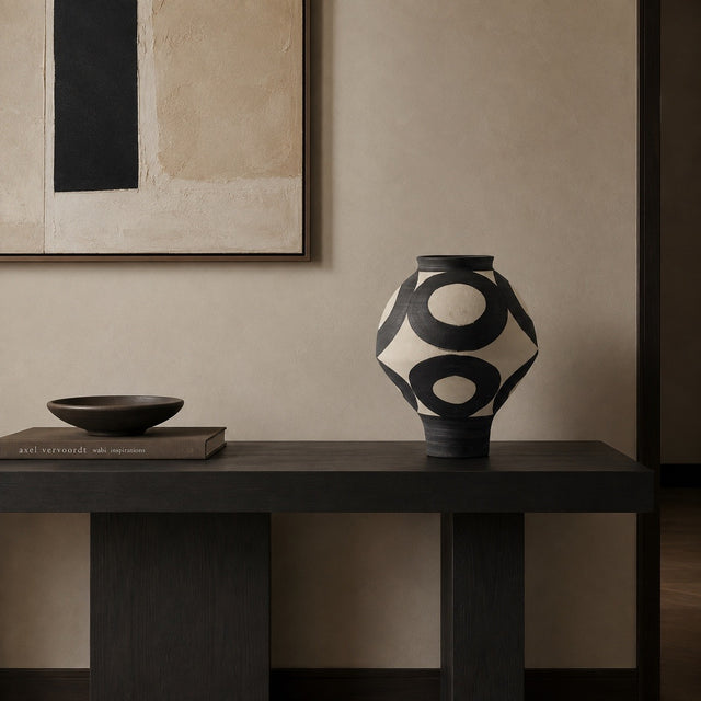 A black and cream geometric patterned ceramic vase sits on a dark wooden console table in a minimalist room setting, next to a small dark bowl and a book.