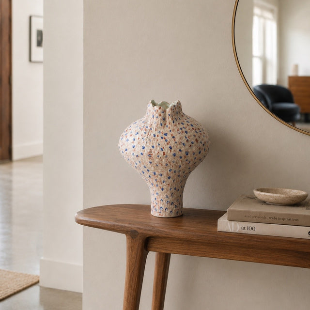 A speckled ceramic vase with a flared rim sits on a wooden console table in a softly lit interior room. Books and a small bowl are stacked on the table, with a mirror and doorway visible in the background.