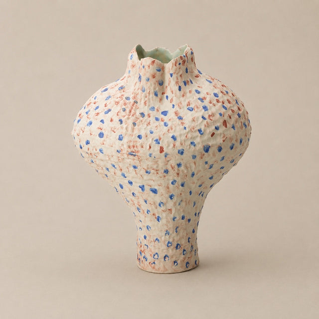 A handmade ceramic vase with a textured, bulbous shape, featuring a speckled pattern of blue and terracotta red dots on a warm cream base, set against a solid beige background.