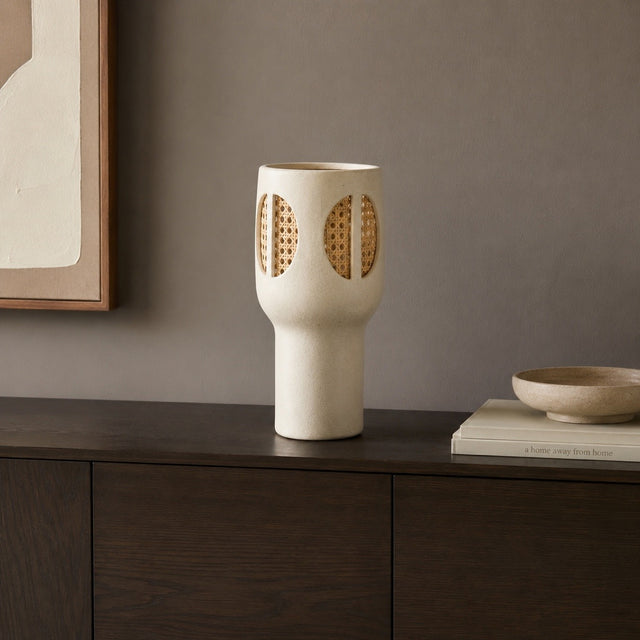 A cream-colored ceramic vase with woven cane accents, displayed on a dark wood console table next to a small bowl and books against a neutral gray wall.