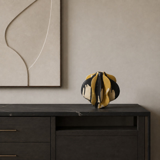 A sculptural, organic-shaped vase with a black and mustard yellow abstract pattern sits on a dark wood sideboard against a beige wall with a framed abstract print.