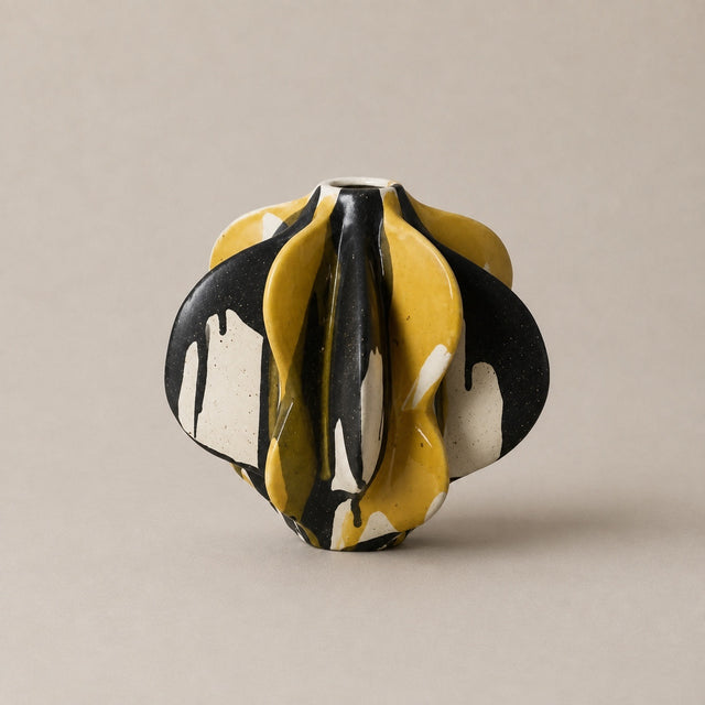 A small sculptural ceramic vase with a fluid, organic dripping pattern in yellow, black, and white, set against a solid warm beige background.