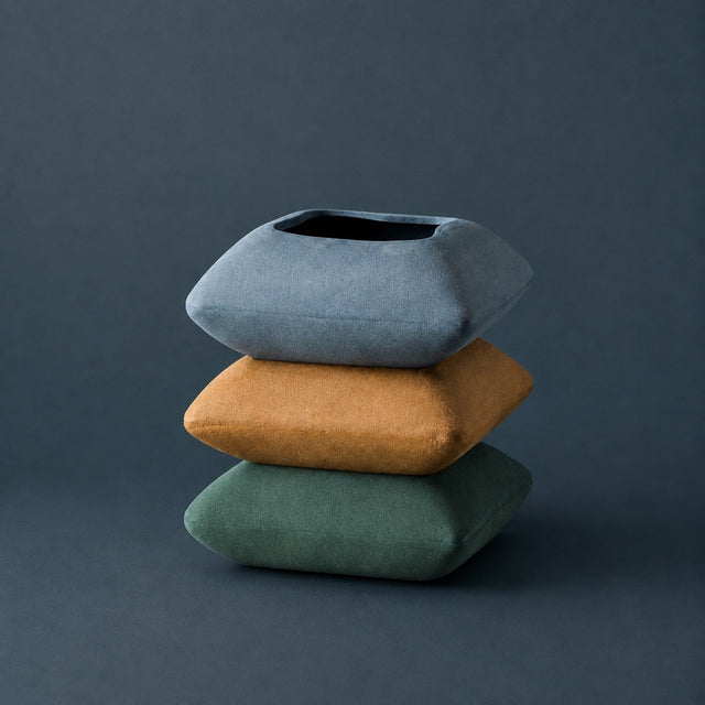Three stacked square decorative cushions in muted blue-gray, mustard yellow, and sage green on a dark slate background.
