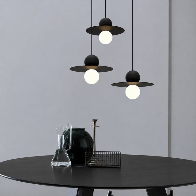 Three modern black pendant lights with wide flat shades and glowing white globe bulbs hang above a round black table in a minimalist interior setting.