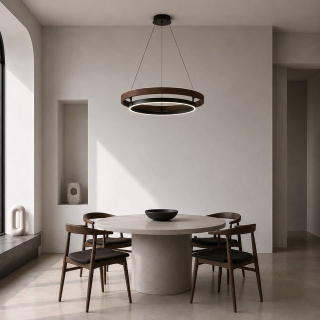 A modern dining room scene featuring a round dining table with four dark wood chairs and a large, circular two-ring pendant light hanging above. The room has minimalist, warm neutral walls and a concrete floor.