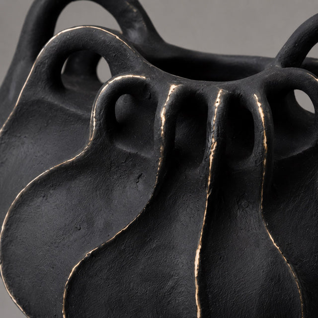 Close-up of a sculptural black ceramic planter with organic wavy contours and a textured matte finish, showing subtle warm metallic edging.