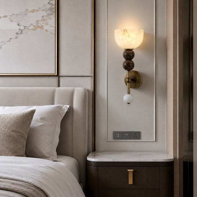 A modern wall-mounted sconce with a glowing frosted glass shade, dark brown spherical accents, and a brass fixture mounted on a beige bedroom wall beside a bed and nightstand.