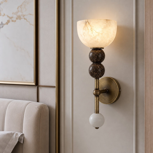 A wall-mounted sconce light fixture with a glowing warm white marble-look bowl shade, three stacked dark brown marbled spherical accents, a brass-toned vertical stem, and a white spherical finial, shown installed on a beige paneled wall next to a sofa.