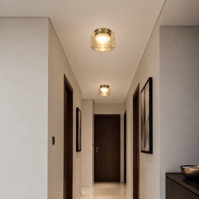 A modern semi-flush mount ceiling light with a brushed gold metal frame and clear glass shade, emitting a warm glow in a hallway with beige walls and dark wood doors.