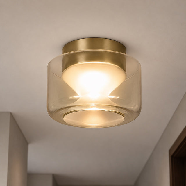 A modern semi-flush mount ceiling light fixture with a brushed brass gold metal top and a clear cylindrical glass shade, emitting a warm yellow glow from the illuminated bulb inside. The fixture is mounted on a white ceiling in an indoor hallway setting.
