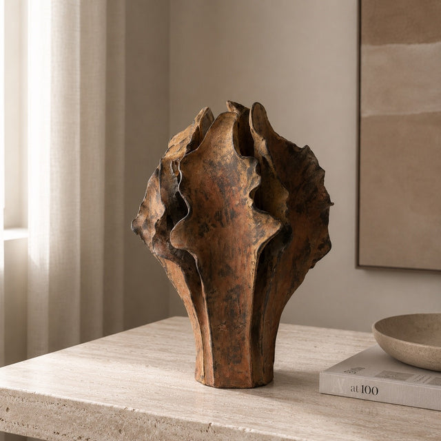 A sculptural rusted metal vase with a textured, organic leaf-like silhouette, displayed on a light stone table in a warm, neutral-toned interior setting.