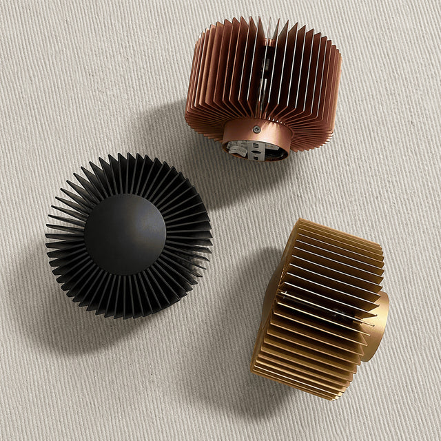 Three ribbed cylindrical heat sinks or finned components arranged on a light textured surface: one matte black, one copper-toned, and one gold-toned.