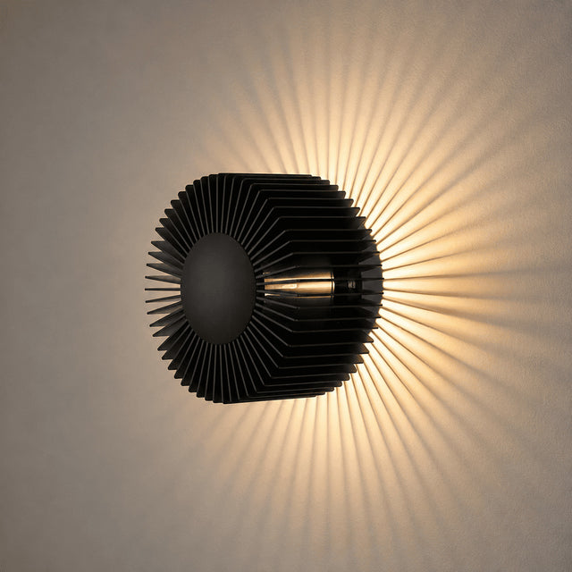 A modern black wall-mounted light fixture with a ribbed, sunburst-like shade casting a warm radial glow on a beige wall.