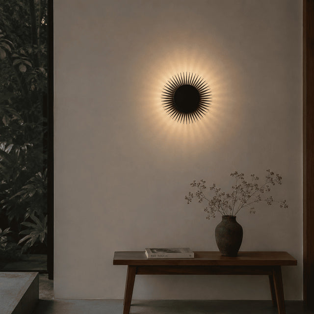 A circular black sunburst wall sconce mounted on a warm off-white wall above a wooden console table with a vase and book, in a styled interior setting.