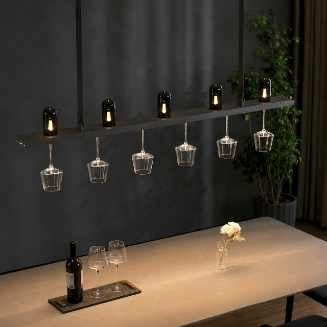 A modern bar pendant light fixture with six black glass bottles on a horizontal bar, each holding a lit candle, with six clear wine glasses hanging below. The fixture is shown in a styled bar or dining environment above a wooden table with a wine bottle and glasses.