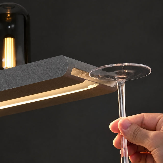 A close-up of a modern under-shelf LED light bar mounted beneath a shelf, illuminating a clear wine glass held by a hand. The background is a dark, minimalist setting with a blurred light bulb.