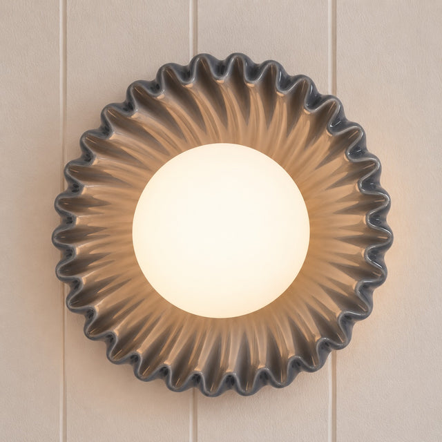 A round wall sconce with a fluted, scalloped outer frame and a glowing frosted white center, mounted on a beige paneled wall.