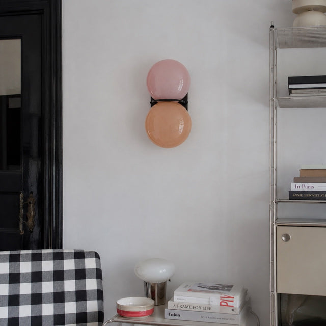 A modern wall-mounted double globe sconce with one pink glass globe and one peach-orange glass globe on a black wall mount, shown in a styled interior setting.