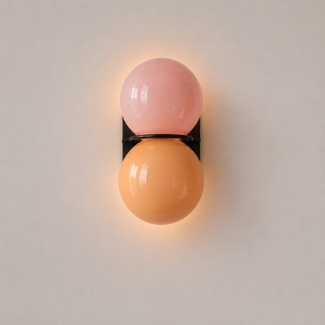 A modern wall-mounted sconce with two glossy spherical shades, one pale pink and one warm orange, separated by a thin black band and mounted on a light gray wall.