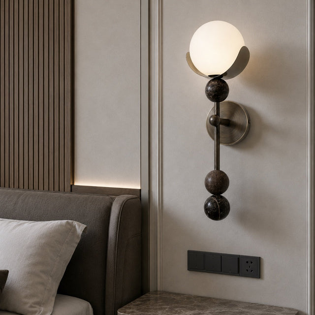 A modern wall-mounted sconce with a glowing white globe shade and dark bronze spherical accents, installed on a beige wall next to a bed.