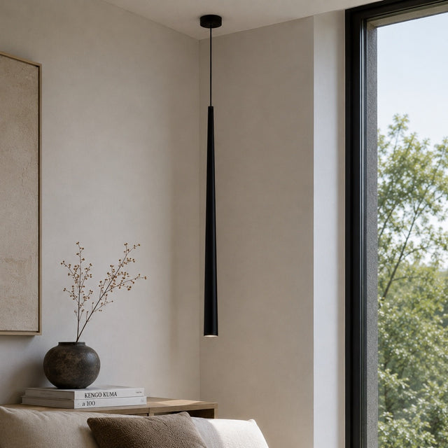 A modern black linear pendant light hanging from a black ceiling mount in a minimalist beige room with a window showing green trees outside.