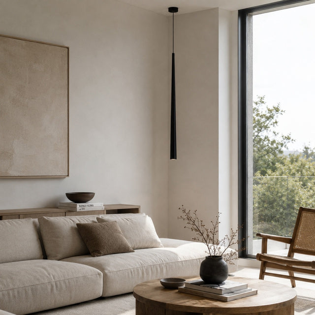 A modern living room corner featuring a light beige sectional sofa, a round wooden coffee table with a black vase, a large framed beige artwork on the wall, and a tall black pendant light. A large window on the right shows green trees outside.