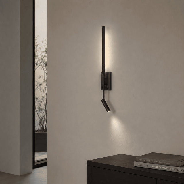 A modern black wall-mounted sconce with a vertical bar and angled side lamp, shown lit on a beige wall above a dark wooden console table.
