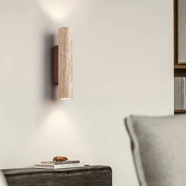 Wall-mounted cylindrical light fixture on a neutral wall with a book and decorative item on a surface below.