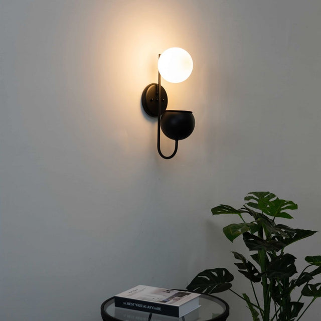 Wall-mounted light fixture on a plain wall with a plant and books in the foreground.