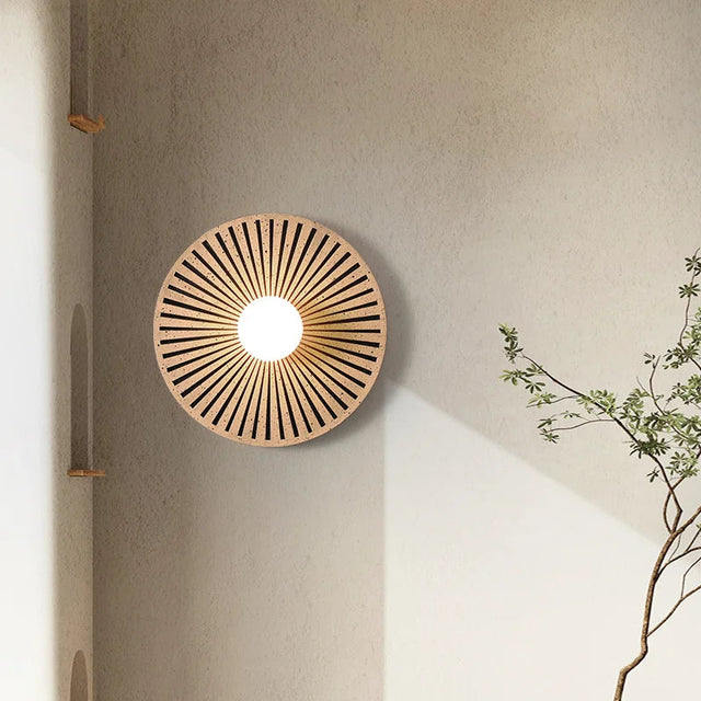 Round wooden wall light fixture on a neutral wall with a plant to the right.