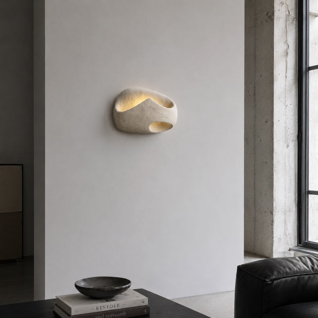 An organic-shaped wall sconce made of textured beige stone or concrete, illuminated from within to cast a warm glow, mounted on a minimalist white wall in a modern interior setting.