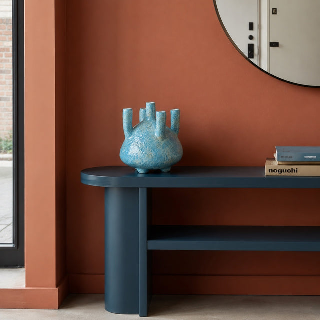 A modern dark blue console table with a cylindrical pedestal base and open shelf, styled with a textured light blue vase and books against a terracotta wall.