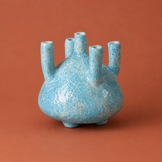 A small ceramic vase with a rounded bulbous body and five short cylindrical necks, covered in a textured light blue glaze, set against a solid terracotta background.