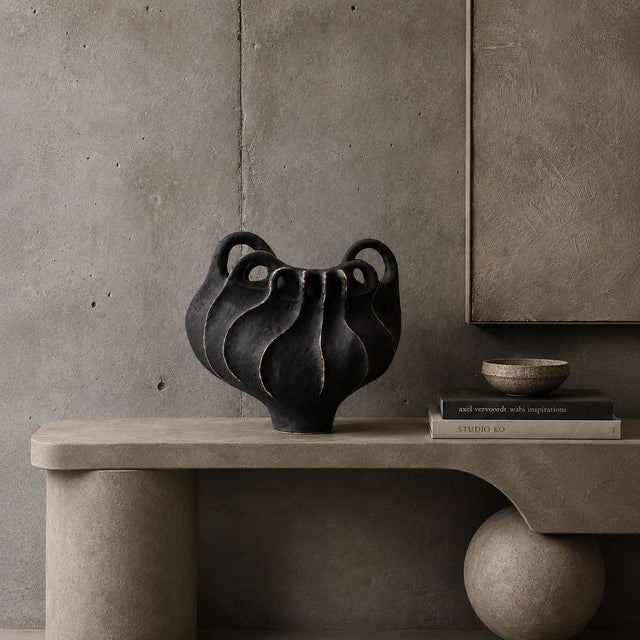 A sculptural black ceramic vase with multiple wavy handles sits on a concrete bench against a textured concrete wall, styled with books and a small bowl.