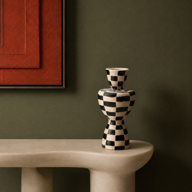 A small decorative vase with a black and cream checkerboard pattern sits on a curved beige stone ledge against a dark olive green wall, with part of a red framed artwork visible at the upper left.