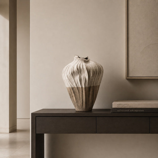 A ribbed ceramic vase in a warm beige and brown glaze sits on a dark wood console table in a minimalist, neutral-toned interior setting.