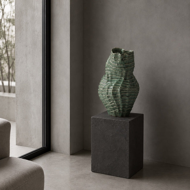 A sculptural, ribbed ceramic vase in a textured sage green glaze sits on a dark charcoal stone pedestal in a minimalist, modern room with concrete walls and a large window.