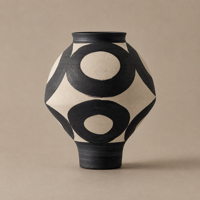A ceramic vase with a matte black and cream geometric pattern featuring bold concentric circles, set against a solid beige background.