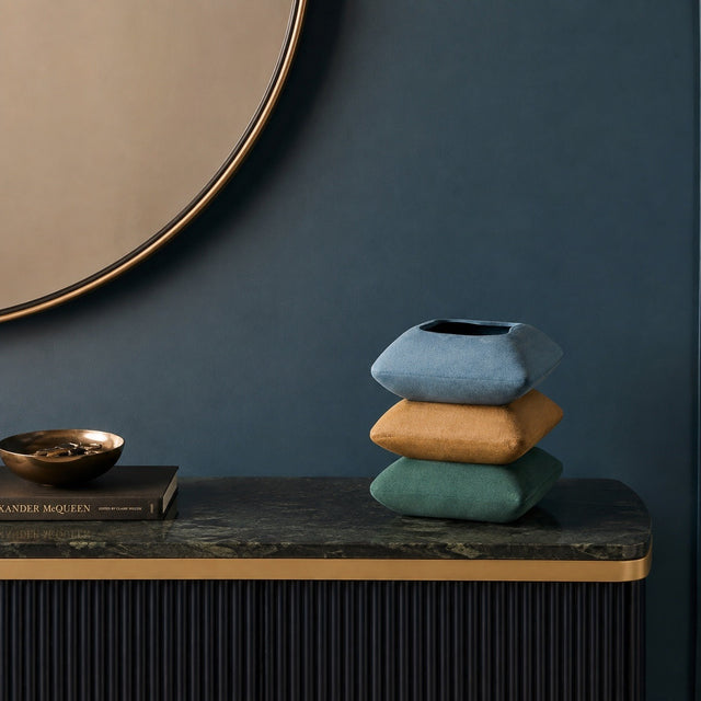 Three square decorative pillows in dusty blue, mustard yellow, and sage green are stacked on a dark marble console table against a dark blue wall.