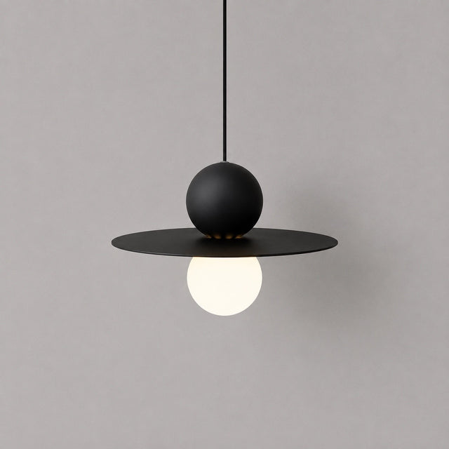 A modern black pendant light with a wide circular disc, a matte black spherical shade, and a glowing white globe bulb, suspended against a plain gray wall.