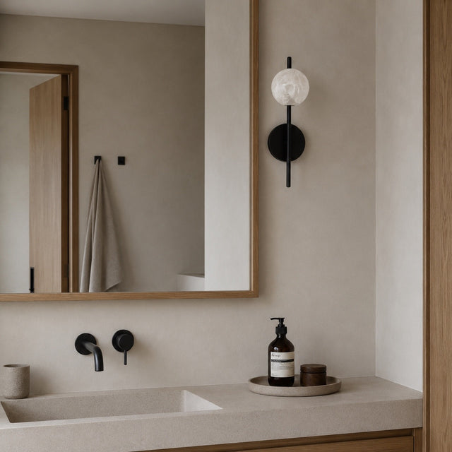 A modern bathroom interior with a beige stone countertop, rectangular sink, wall-mounted mirror with a wood frame, black faucet controls, a black wall sconce with a white globe bulb, and a soap dispenser and small jar on a tray.