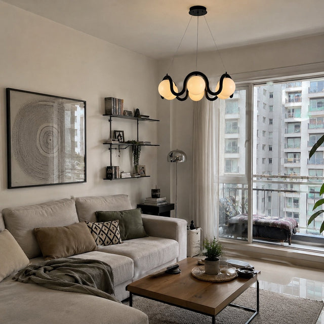 A modern living room featuring a beige sectional sofa with decorative pillows, a wooden coffee table, a black wavy pendant light, and a large framed circular print on the wall. The room is well-lit with natural light coming from a large window overlooking other apartment buildings.