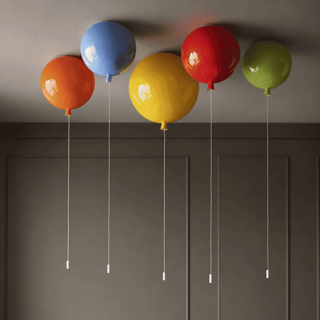 Five colorful balloon-shaped pendant lights in orange, blue, yellow, red, and green, suspended from the ceiling by thin white cords against a dark gray paneled wall.