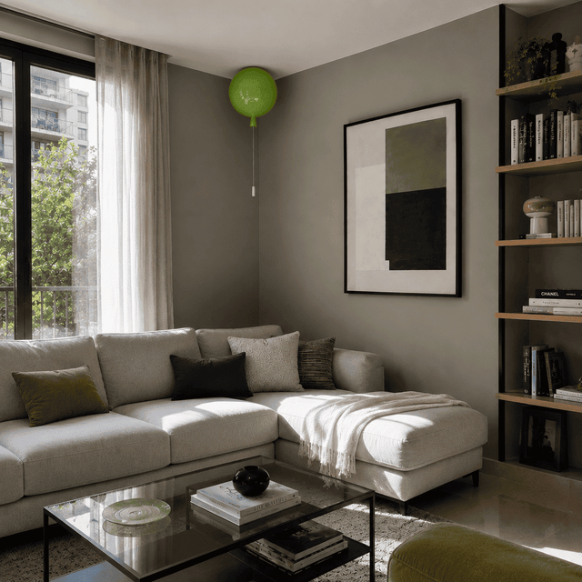 A modern living room featuring a light beige sectional sofa with accent pillows, a glass-top coffee table, a bookshelf on the right, and a gray wall with framed abstract art. A bright green balloon floats near the top center.