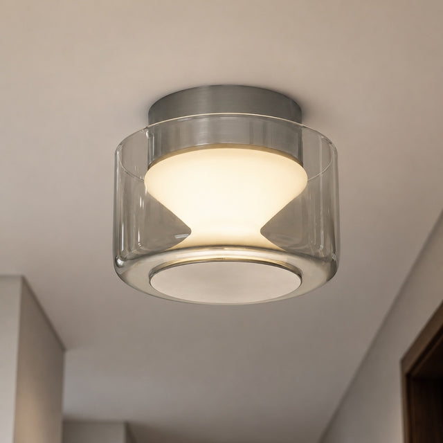 A modern semi-flush mount ceiling light with a clear glass cylinder shade, brushed silver metal trim, and a warm white LED light glowing inside, mounted on a white ceiling.