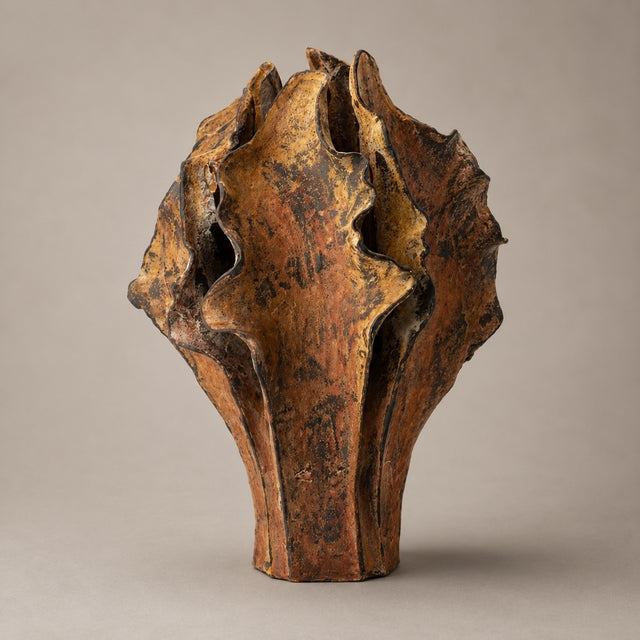 An abstract sculptural ceramic vase with organic, layered organic shapes, featuring a textured glaze in earthy brown, rust-orange, and black tones, set against a solid warm gray background.