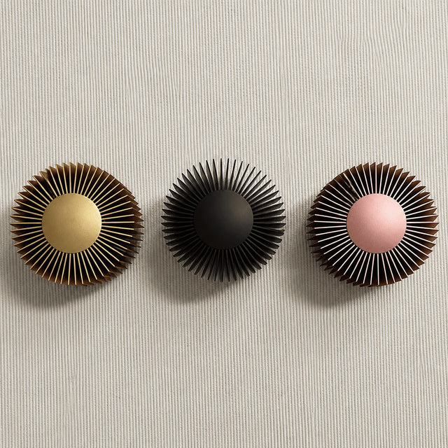 Three round decorative wall-mounted pieces with radiating slats are arranged horizontally on a light textured wall. The left piece has a gold center, the middle piece is black, and the right piece has a pink center.