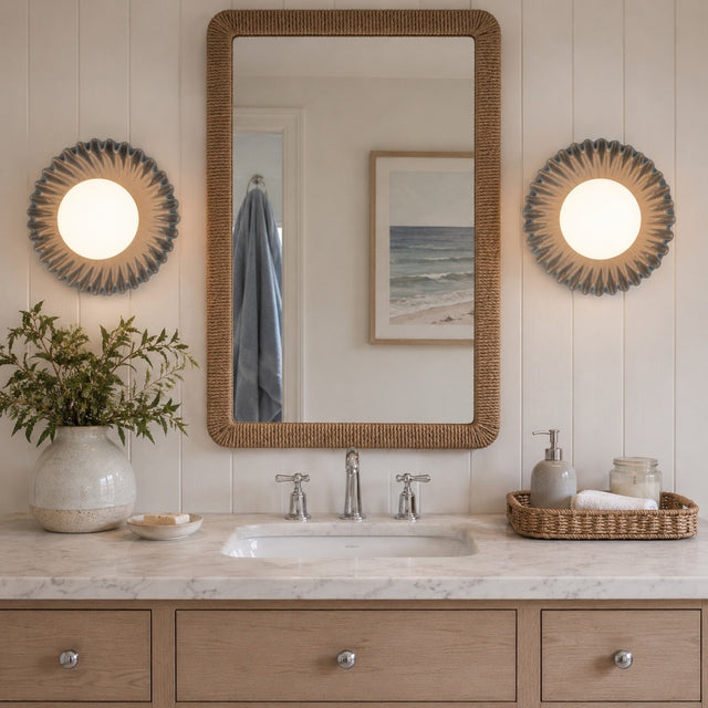 A styled bathroom vanity scene with a large rectangular woven mirror centered above a marble sink, flanked by two round sunburst wall lights. A vase with greenery, a soap dish, and a tray with toiletries sit on the counter against a light wood cabinet and paneled wall.